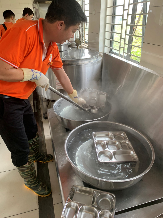A person in an orange shirt and gloves stirring a pan with steam

Description automatically generated